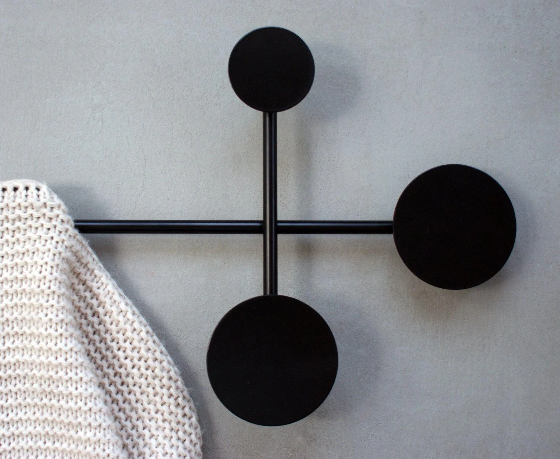 A modern wall hanger with black, round hooks, mounted on simple, thin rods. A cream, knitted fabric hangs on the hook. The background is a smooth, gray wall.