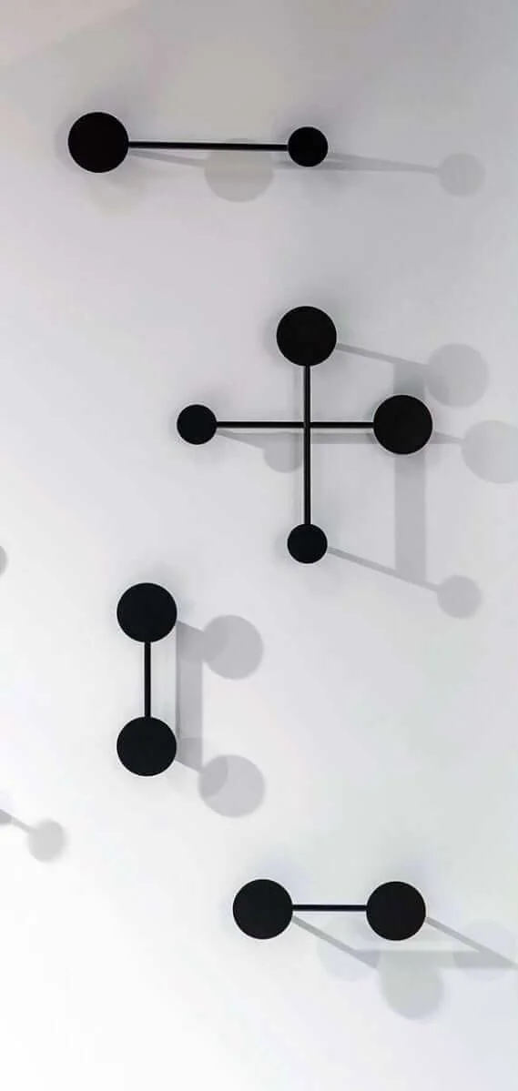 A minimalist, modern wall hanger consisting of black, round hooks and thin rods, mounted on a smooth, white wall. It creates geometric patterns, and the shadows of the hooks emphasize its simple form.