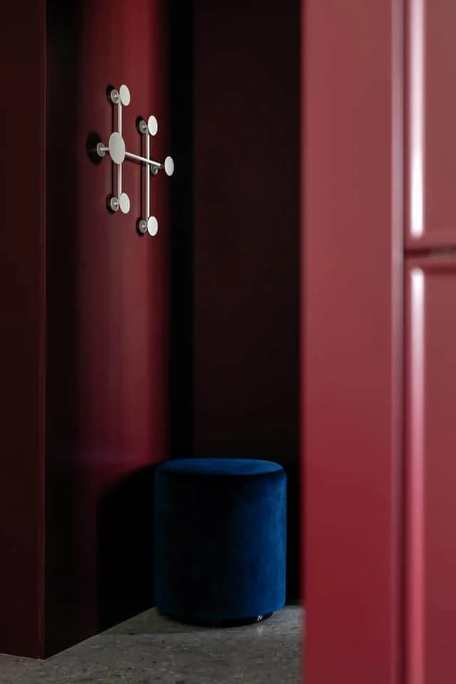 A wall hanger with white, round hooks, mounted on a dark red wall. No items hang on the hook, and a navy upholstered pouf is visible in the background.