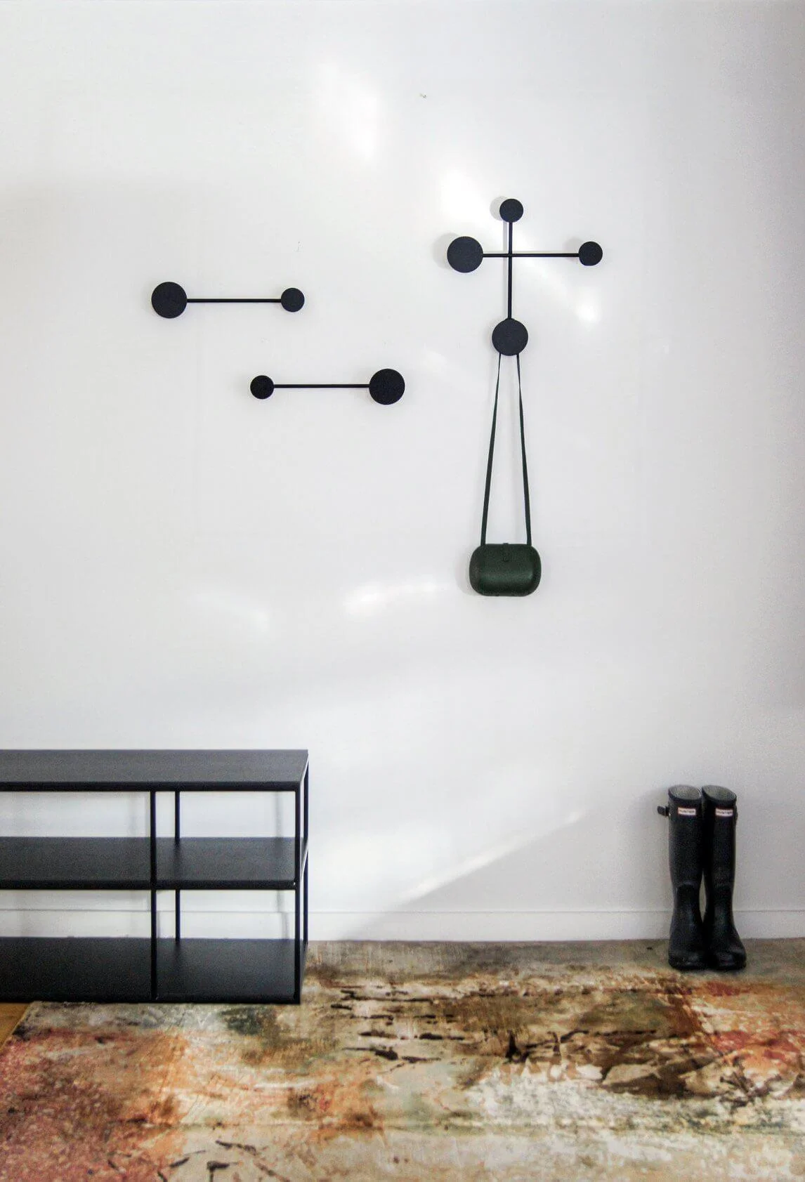 A black, modern wall hanger in the form of circles and lines, with a small green handbag hanging on it. The background features a white, minimalist wall, a black shelf, and a pair of black rain boots.