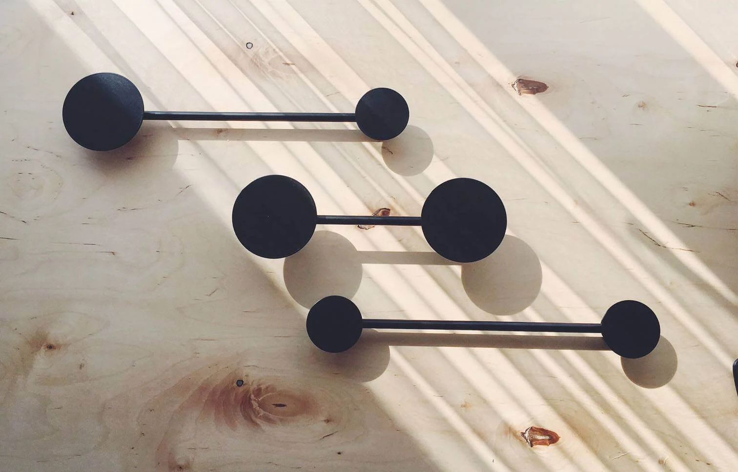 A modern, minimalist wall hanger with black, round hooks, mounted on a light wooden surface, with angled light rays creating distinct shadows.