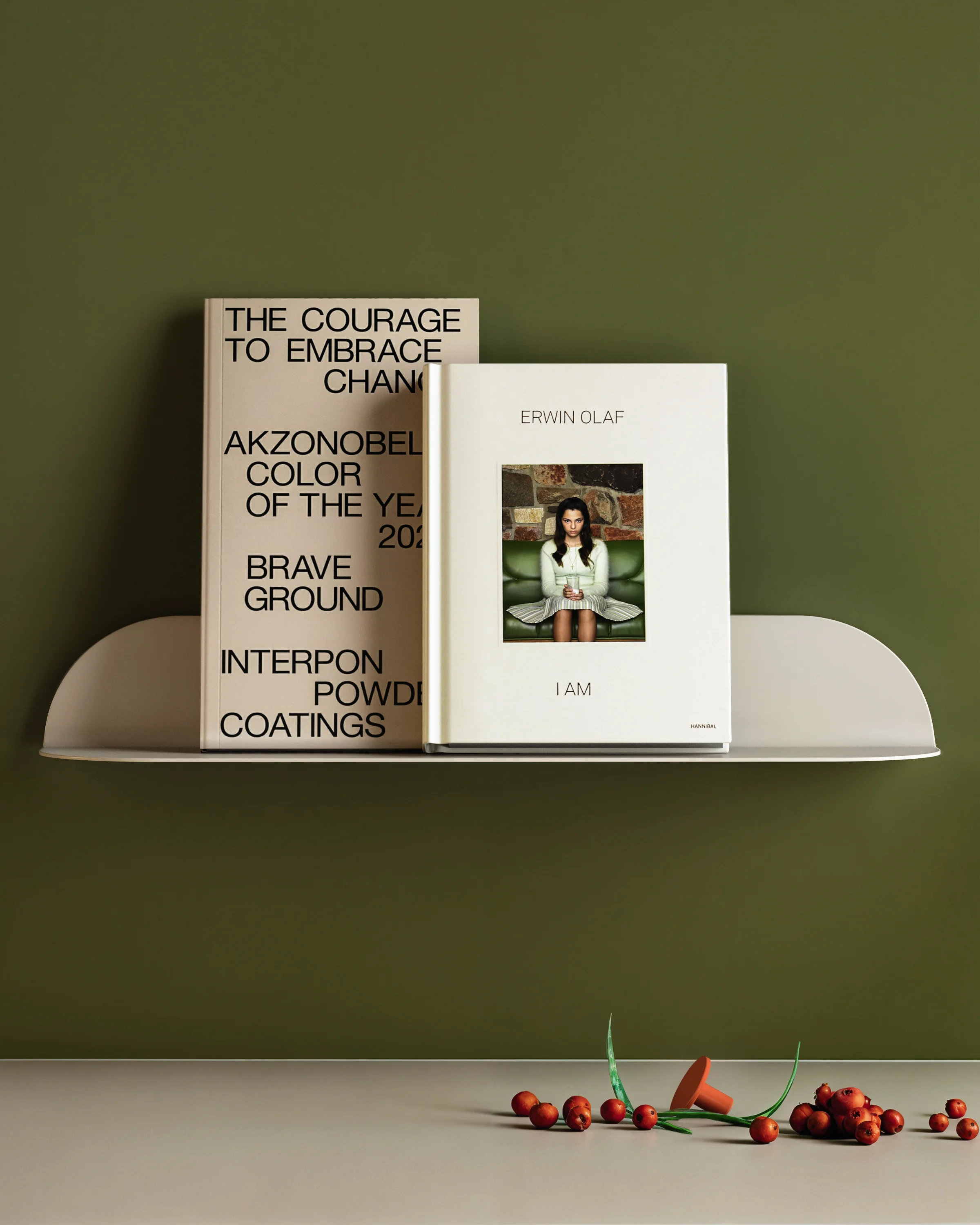 Two books on a white shelf against an olive-green wall. One book features text on a light cover, the other a photograph of a woman. Below the shelf, red fruits and green stems rest on the surface.