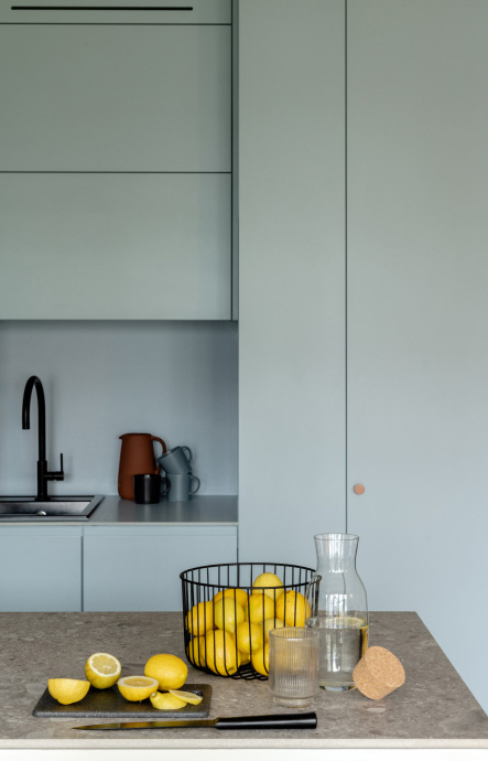 In the background there is a set of kitchen furniture in light blue color, with round, orange handles. In a niche there is a pitcher with mugs, to their left there is a sink. In the foreground there is a countertop on which lies a bowl with lemons, a carafe with water, and a cutting board with a knife. On the board there are some cut lemon halves.