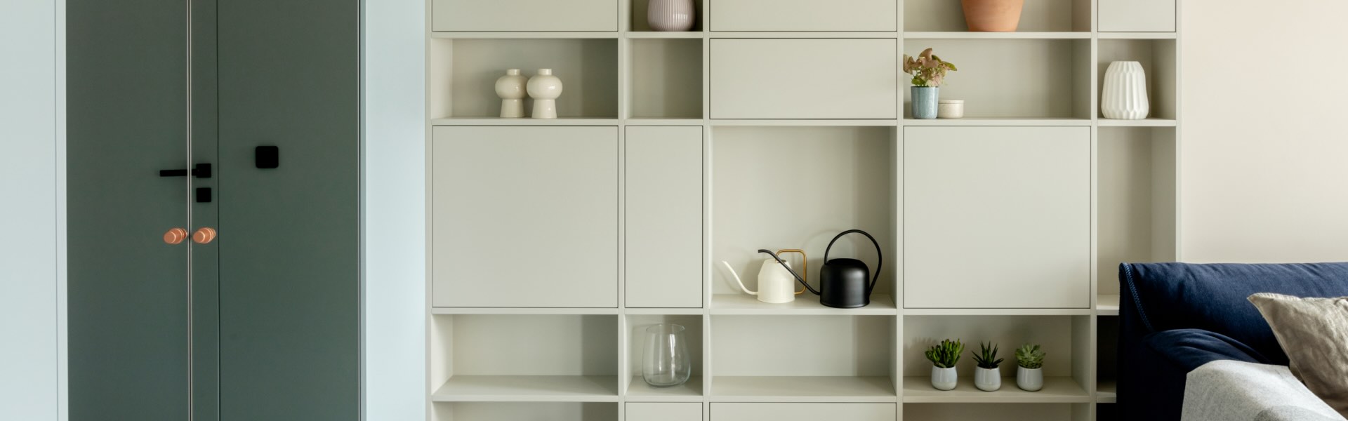 A room with a white set of cabinets arranged in a checkerboard pattern. Closed cabinets with white fronts are handleless, on the open shelves lie vases, pots, a glass, and small plants. On the left side there is a cabinet with mirrored doors and orange, round handles. On the right side there is a blue sofa with pillows.