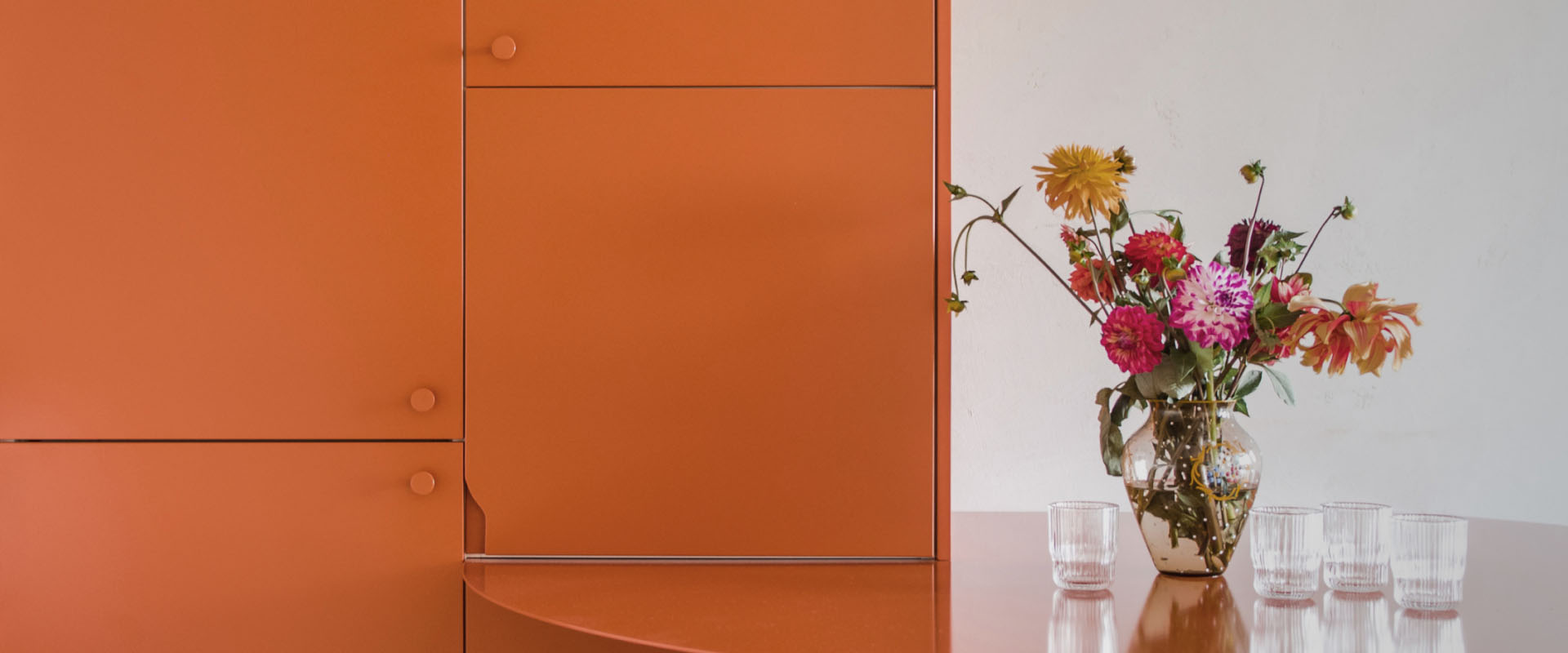 Komplet pomarańczowej szafy i stołu w tym samym kolorze na tle białej ściany. Przed stołem stoi drewniane krzesło, na stole stoi szklanka, z prawej strony cięte polne kwiaty.